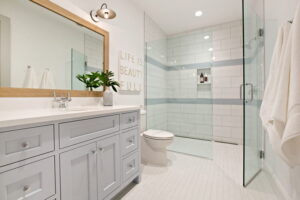 a photo of a bathroom that shows a full-size mirror above the sink with gray cupboards. The toilet is next to the sink between the standing shower. The mirror is rimmed in gold and there's a plan on the counter.