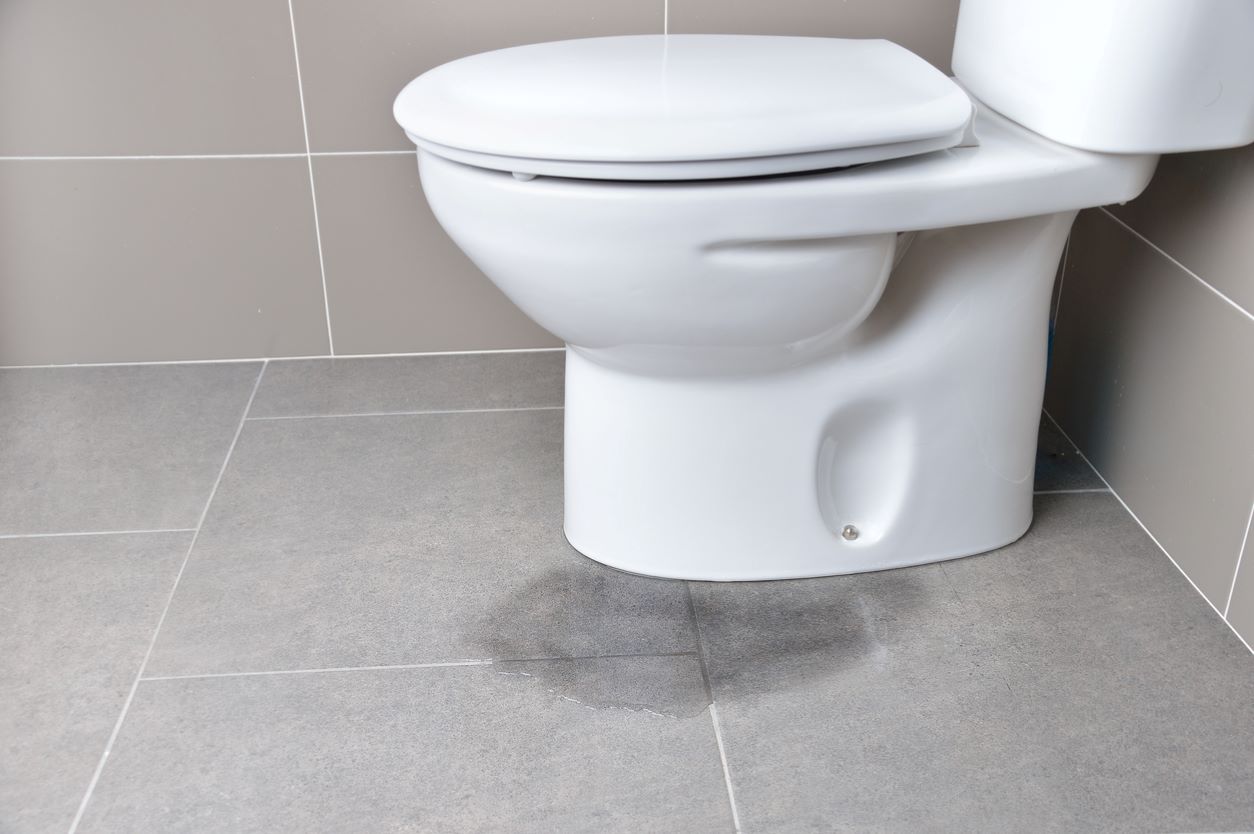 White toilet with small leak of water at base of toilet pooling up on gray tile floor