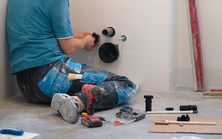 A plumber with power tools surrounding him as he works on pipes inside of a wall.